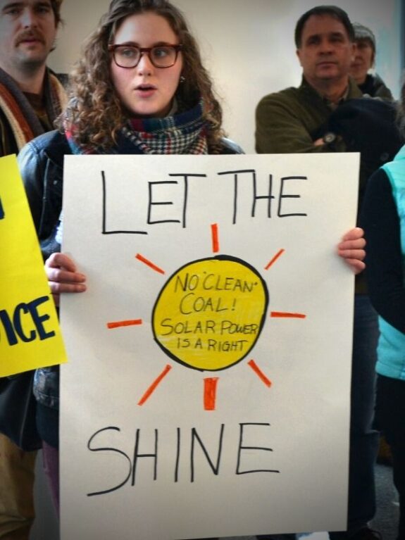 A woman protesting at the Minnesota State Capitol holding a sign that says "Let the Sun Shine - No Clean Coal! Solar Power is a Right!"